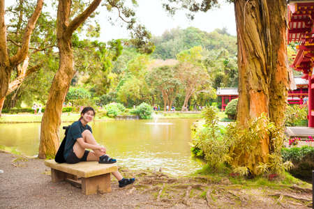 Biracial teen girl resting on concrete bench next to quiet Asian style pond in an Asian temple gardenの写真素材
