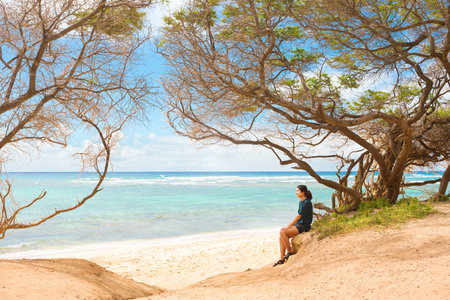 One biracial teen girl sitting alone on empty beach under windswept trees on beach along blue ocean  on tropical island