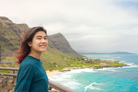 Biracial  teen girl standing  above tropical Hawaiian scenic view of blue ocean and green mountains on Oahu, Hawaiiの写真素材