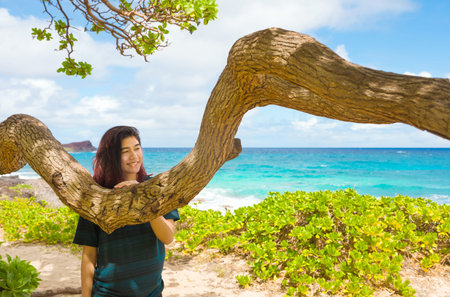 Biracial teen girl under a gnarly shade tree by blue Hawaiian ocean