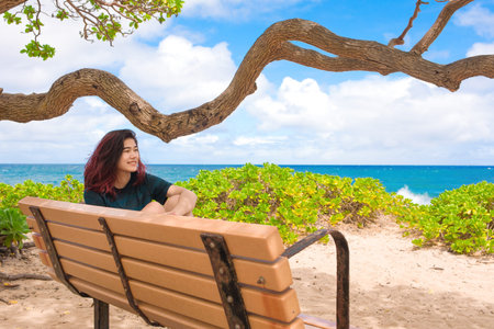 Biracial teen girl relaxing on a bench by Hawaiian ocean under gnarly shade tree
