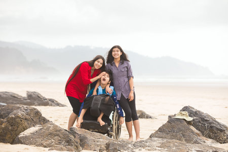 Two biracial  teen sisters hugging disabled little brother in wheelchair on beach by ocean on foggy...
