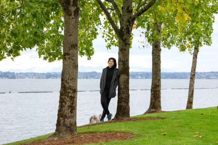 Smiling biracial  teen girl or young adult female in gray jacket leaning against tree in park by lakeの写真素材
