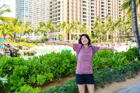 Young woman excited about trip in Hawaii, arms outstretched with resort building in backgroundの写真素材
