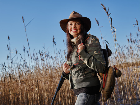 Waterfowl hunting, smiling female hunter carry a shotgun and a decoys, reeds and blue sky on backgroundの写真素材