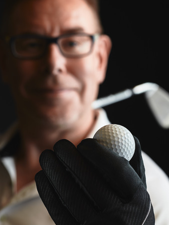 Close up, mature man golfer wearing a white shirt and he holds a golf ball on hand, iron golf club on his shoulder - focus on ball, studio shot, black backgroundの写真素材