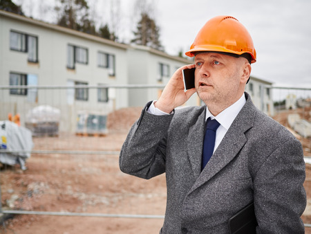 Male engineer uses cell phone, he wearing the suit and the hard hat, house construction site on backgroundの写真素材