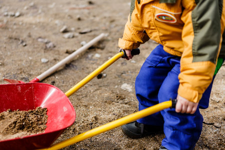 child in blue work pants play with sand, red shovel and small barrow