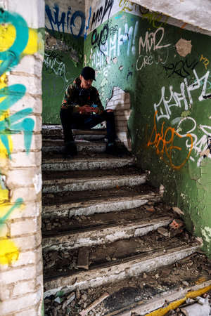 Young teenage boy sitting on stairs in ruined building and using smarthphone.の写真素材