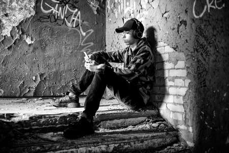 Young teenage boy with headphones sitting on stairs in ruined building using smarthphone and listening music.の写真素材