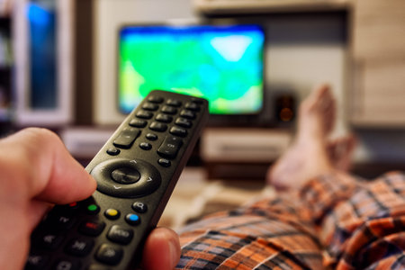 Person lying on bed with crossed legs and switching television channels on TV with finger on remote controllerの写真素材