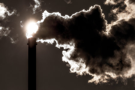 Dark smoke and steam from chimney in factory against the sun on sky