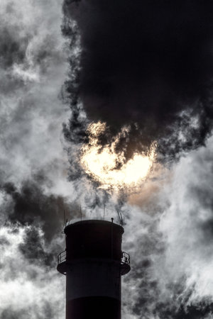 Dark smoke and steam from chimney in factory against the sun on sky