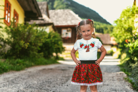 Happy small four years old girl dressed in red folk skirt posing in traditional rural village Vlkolinec with wooden cottages in Slovakiaの写真素材