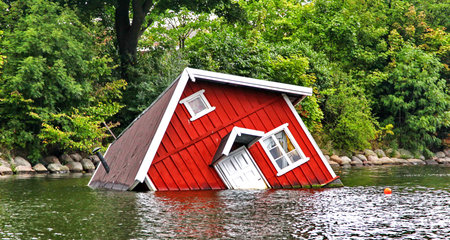 Red wooden house sinking in flood water among green trees, symbol of natural disaster, climate change and environmental destructionの写真素材