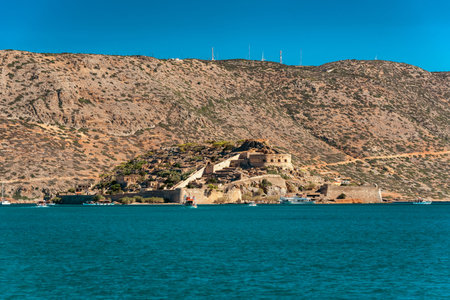 Scenic view of Spinalonga island fortress in Crete, Greece, surrounded by turquoise sea with tourist boats and rocky hills in the background, popular travel and holiday destination in Mediterranean.の写真素材