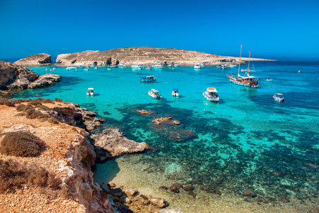 Blue Lagoon with boats anchored in clear turquoise water at Comino island, Malta, with rocky coast and bright summer sky, famous Mediterranean travel destination.の写真素材