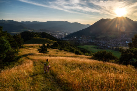 Mother with child walking on hiking trail in golden meadow at sunset above Ruzomberok Slovakia, scenic summer landscape with mountains, town and sun rays in evening sky.の写真素材