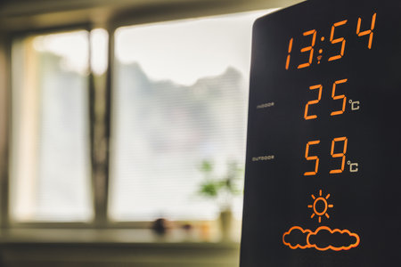 Modern digital weather station showing indoor temperature and humidity on display during hot summer day at home with bright sunlight through window.の写真素材