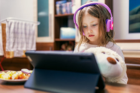 Girl using tablet with headphones while sitting at table in living room. Concept of remote education, entertainment and excessive screen time among children.の写真素材