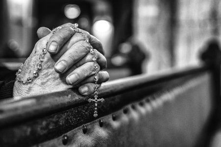 Black and white close up of elderly hands clasped in prayer with rosary beads in church pew. Concept of faith, devotion and spirituality.の写真素材
