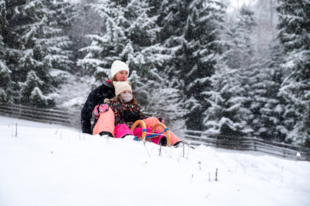 Mother and daughter riding a sled down a snowy hill, enjoying active winter fun together. Family bonding, snowfall, outdoor recreation and joyful seasonal activity in fresh snow.の写真素材