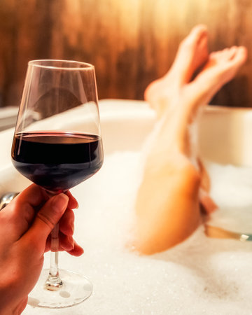 Glass of red wine held above a foam filled bathtub during a relaxing evening with warm light and a calm intimate atmosphere.の写真素材