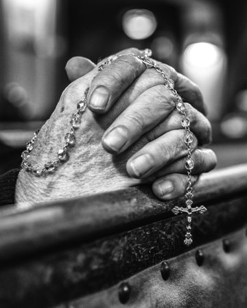 Black and white closeup of hands holding a rosary during prayer inside a quiet church with dramatic soft light.の写真素材