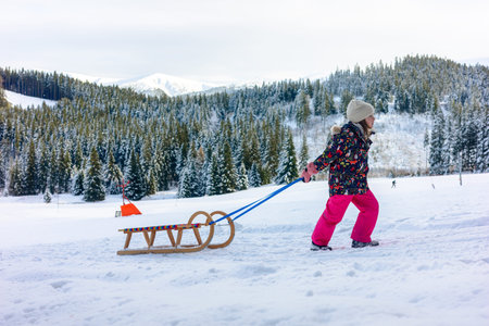 Child pulling wooden sled through fresh snow with forested mountains in background. Winter outdoor...