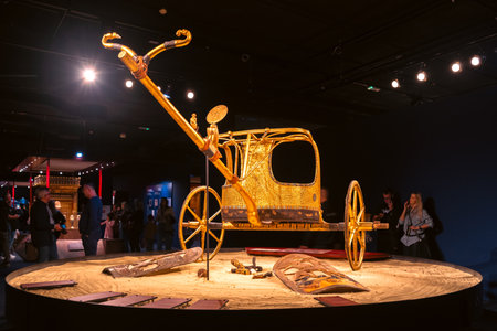 Bratislava, Slovakia - November 15, 2025: Golden ceremonial chariot inspired by ancient Egyptian royal processions displayed at the Tutankhamun exhibition, with visitors observing the artifact in the museum hall.のeditorial素材