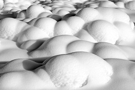 Rounded snowdrifts forming organic shapes under strong winter light. Deep shadows emphasize texture and relief, creating minimalist abstract winter background with high contrast.の写真素材