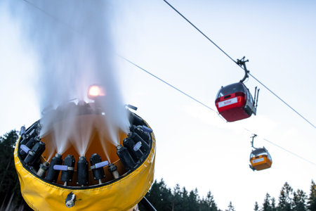 Malino Brdo, Slovakia - December 29, 2025: Snowmaking machine spraying artificial snow while cable cars operate above ski slope during winter season preparation in mountain resort.のeditorial素材