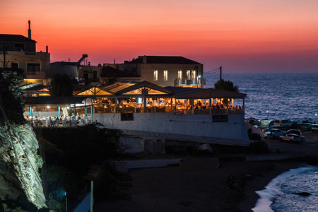 Panormos, Crete, Greece - September 6, 2025: Seaside restaurants and small harbor at sunset with warm lights reflecting on the water in coastal village of Panormos.のeditorial素材