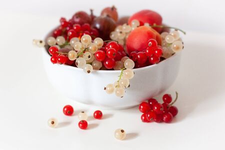White bowl full of red and white currants, gooseberries and apricots on a white table.の写真素材