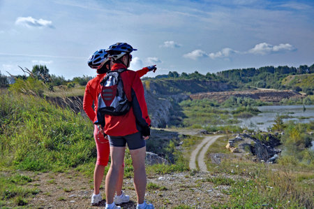 Two cyclists in helmets and red jackets stopped on the edge of the quarry, against the background of a slightly cloudy sky and show something in the distance.の写真素材
