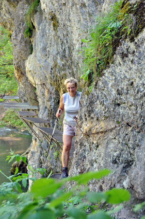 A tourist in a light gray shirt and with a trekking stick, smiling wanders along the river canyon, walking on the platforms mounted in the rock and sticking to the chain.の写真素材