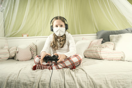 Girl in a mask playing on a game console during quarantine on the bed.