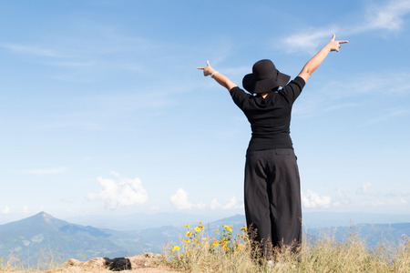 Happy young asia woman enjoying sunny day. black dress of woman standing in meadow with mountain background, freedom in summerの写真素材