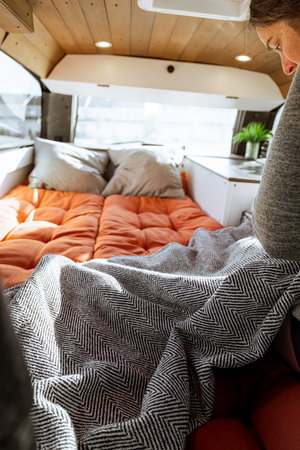 Young 30s caucasian woman with dark hair making the bed in a self converted minivan campervan for van life with lovely light coming in through the windows.の写真素材