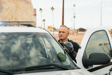 Horizontal image of white male caucasian police officer standing in the door opening of his trooper car with lights on looking away from camera next to railroad tracks.の写真素材