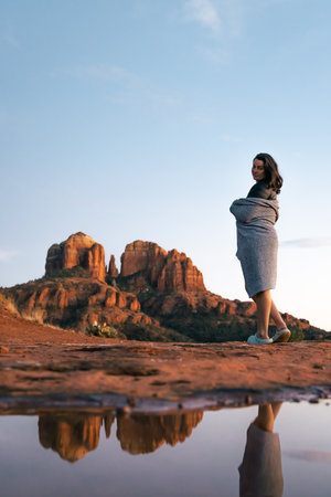 Young good looking female woman standing with blanket wrapped around her looking at camera with red rock cathedral rock butte lit up by incredible sunset colors during blue hour in Sedona Arizona USA.の写真素材