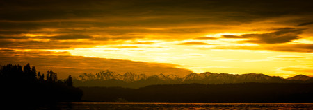 A panorama shot on the Pacific northwest with water in the foreground, an evergreen forest in the middle of the image with distant snow covered mountains.  The clouds in the sky are brilliantly lit from below.の写真素材
