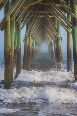 A view under a pier at a beach with rough seas and some light fog as the viewer looks through...