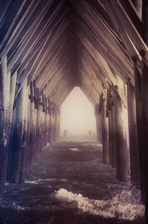 A view under a pier at a beach with rough seas and some light fog as the viewer looks through pillars to the open ocean at the other end.の写真素材