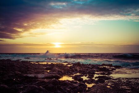 The sun rising over the ocean with a rocky foreground and rough surface crashing into the shore of Hawaii.の写真素材