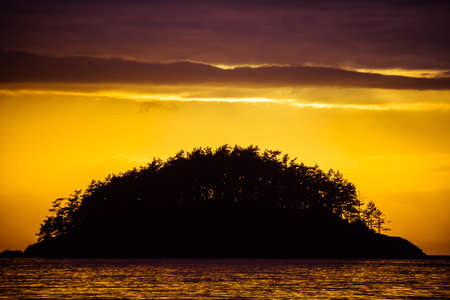 Small island in silhouette against a brilliant sunset and lots of colorful reflections in the water.の写真素材