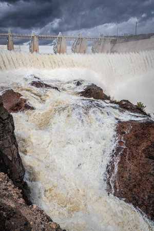 A dam with its flood gates open and a large volume of water flowing over rocks with storm clouds.の写真素材