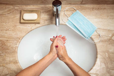 Young woman washing and rubbing her wet hands with soap, two new surgical masks ready on her right during pandemiaの写真素材