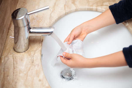 Young kid washing his cloth mask and rinsing off soap under tap water during pandemia with rolled-up...