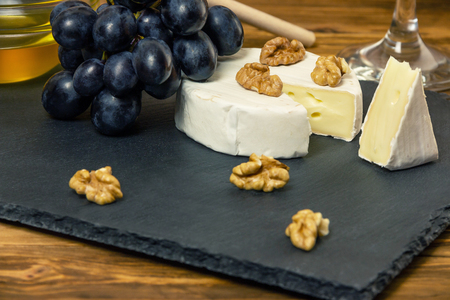 Camembert cheese in a dark tray next to sweet blue grapes nuts and honey on wooden backgroundの写真素材